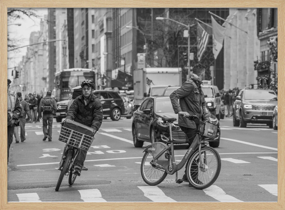 A black and white street photograph of a busy city intersection, framed in silver. In the foreground, two men are crossing with their bicycles amidst traffic, with one cyclist looking directly at the camera. Tall buildings and other pedestrians line the background, capturing a moment of everyday city life. Artwork