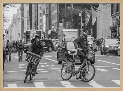A black and white street photograph of a busy city intersection, framed in silver. In the foreground, two men are crossing with their bicycles amidst traffic, with one cyclist looking directly at the camera. Tall buildings and other pedestrians line the background, capturing a moment of everyday city life. Artwork