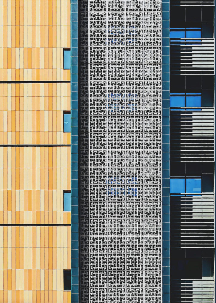 A vertical abstract architectural photograph showcasing the facade of a modern building, featuring a contrast of textures and colors: warm yellow vertical tiles on the left, an intricate white geometric screen in the center, and a dark section with vibrant blue windows on the right. Print