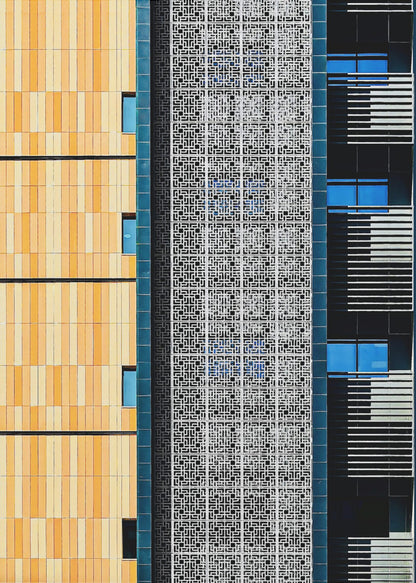 A vertical abstract architectural photograph showcasing the facade of a modern building, featuring a contrast of textures and colors: warm yellow vertical tiles on the left, an intricate white geometric screen in the center, and a dark section with vibrant blue windows on the right. Print