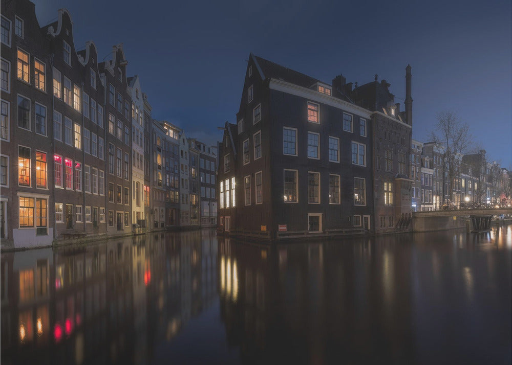 A framed photograph capturing a serene night view of an Amsterdam canal. Traditional Dutch houses with lit windows line the calm water, casting warm reflections under a deep blue twilight sky. Wall Art