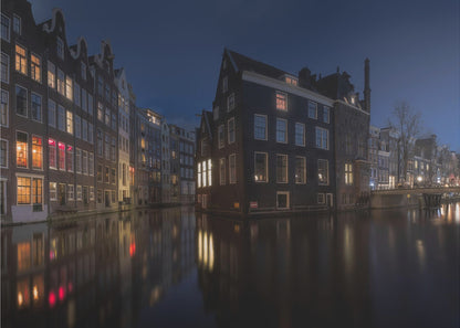 A framed photograph capturing a serene night view of an Amsterdam canal. Traditional Dutch houses with lit windows line the calm water, casting warm reflections under a deep blue twilight sky. Wall Art