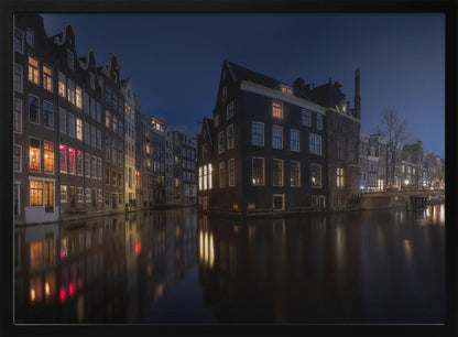 A framed photograph capturing a serene night view of an Amsterdam canal. Traditional Dutch houses with lit windows line the calm water, casting warm reflections under a deep blue twilight sky. Wall Art