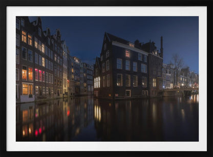 A framed photograph capturing a serene night view of an Amsterdam canal. Traditional Dutch houses with lit windows line the calm water, casting warm reflections under a deep blue twilight sky. Wall Art