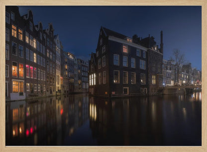 A framed photograph capturing a serene night view of an Amsterdam canal. Traditional Dutch houses with lit windows line the calm water, casting warm reflections under a deep blue twilight sky. Wall Art