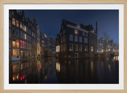 A framed photograph capturing a serene night view of an Amsterdam canal. Traditional Dutch houses with lit windows line the calm water, casting warm reflections under a deep blue twilight sky. Wall Art