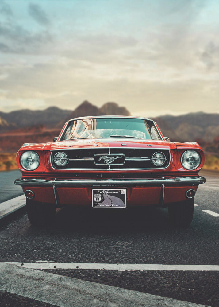 A low-angle, head-on shot of a vintage red Ford Mustang parked on an asphalt road. The iconic silver horse emblem is visible on the grille. In the background, a desert mountain range is blurred under a cloudy, muted sky. Poster