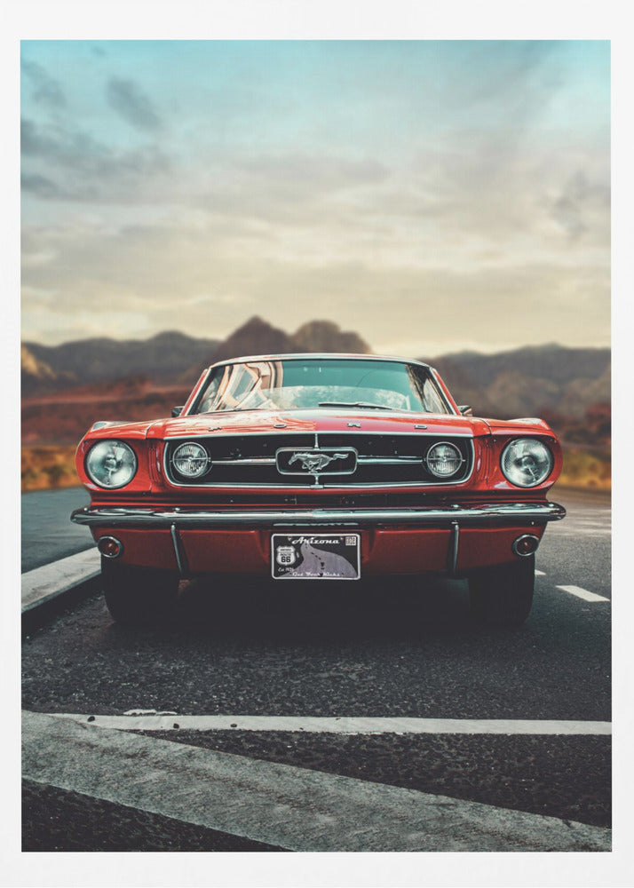 A low-angle, head-on shot of a vintage red Ford Mustang parked on an asphalt road. The iconic silver horse emblem is visible on the grille. In the background, a desert mountain range is blurred under a cloudy, muted sky. Poster