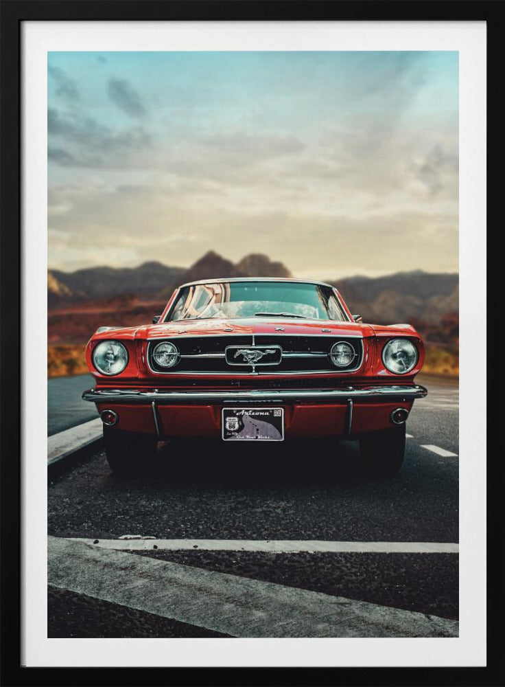 A low-angle, head-on shot of a vintage red Ford Mustang parked on an asphalt road. The iconic silver horse emblem is visible on the grille. In the background, a desert mountain range is blurred under a cloudy, muted sky. Poster