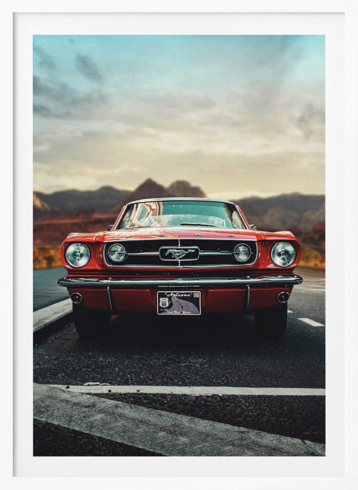 A low-angle, head-on shot of a vintage red Ford Mustang parked on an asphalt road. The iconic silver horse emblem is visible on the grille. In the background, a desert mountain range is blurred under a cloudy, muted sky. Poster