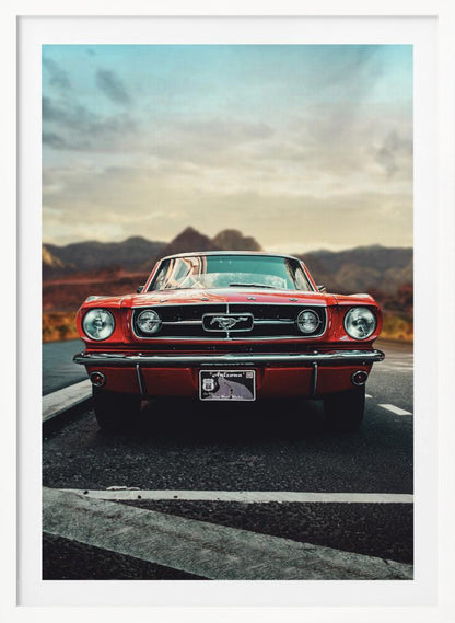 A low-angle, head-on shot of a vintage red Ford Mustang parked on an asphalt road. The iconic silver horse emblem is visible on the grille. In the background, a desert mountain range is blurred under a cloudy, muted sky. Poster