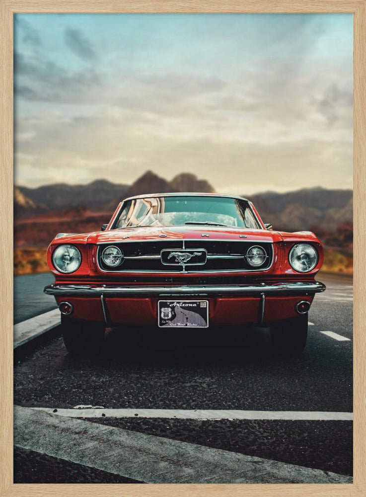 A low-angle, head-on shot of a vintage red Ford Mustang parked on an asphalt road. The iconic silver horse emblem is visible on the grille. In the background, a desert mountain range is blurred under a cloudy, muted sky. Poster