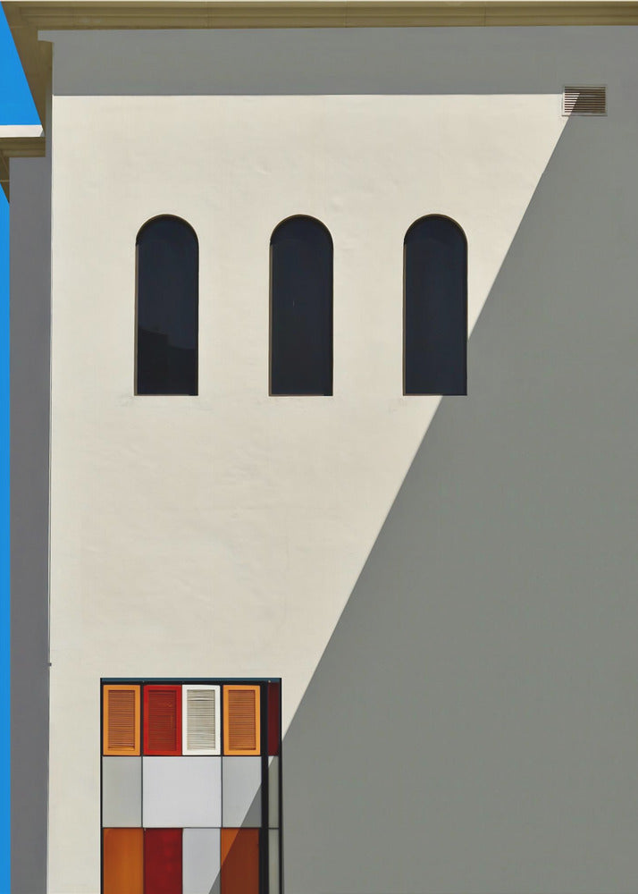 A minimalist photograph of a cream-colored building facade with a sharp diagonal shadow. Three black arched windows are arranged horizontally, and below them, a colorful grid of red, orange, and white panels. A vibrant blue vertical stripe is visible on the far left edge. Wall Art