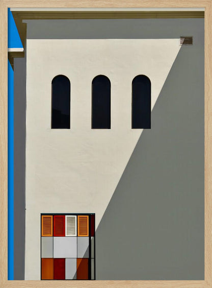 A minimalist photograph of a cream-colored building facade with a sharp diagonal shadow. Three black arched windows are arranged horizontally, and below them, a colorful grid of red, orange, and white panels. A vibrant blue vertical stripe is visible on the far left edge. Wall Art
