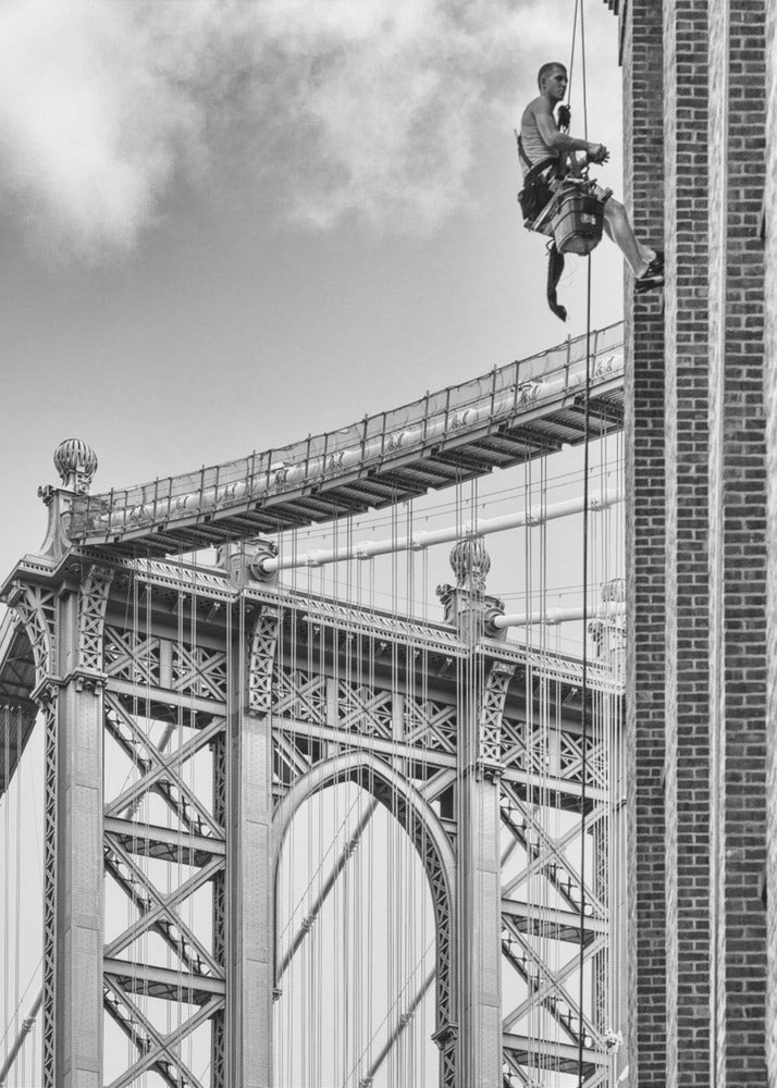 A black and white photograph capturing a window washer rappelling down the side of a tall brick building. In the background, the intricate steel structure of the Manhattan Bridge stands against a partly cloudy sky, creating a powerful urban scene. Decor