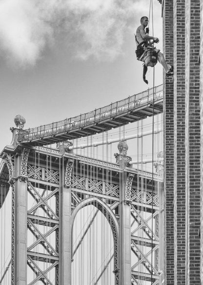 A black and white photograph capturing a window washer rappelling down the side of a tall brick building. In the background, the intricate steel structure of the Manhattan Bridge stands against a partly cloudy sky, creating a powerful urban scene. Decor