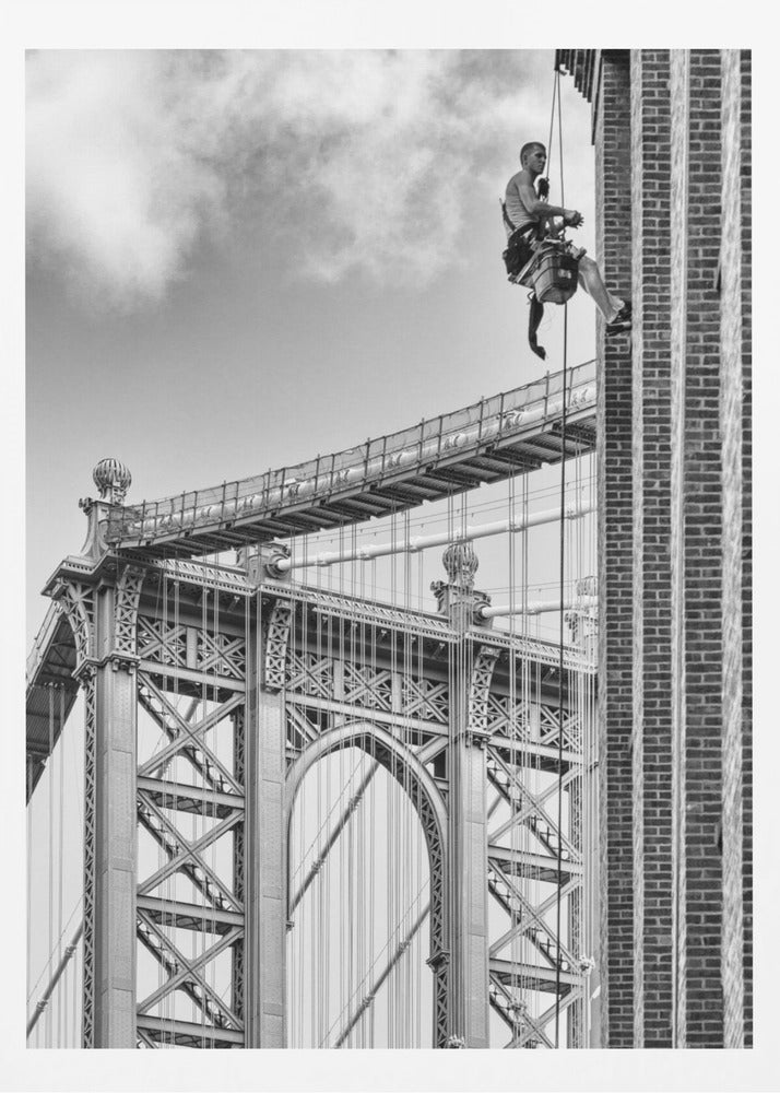 A black and white photograph capturing a window washer rappelling down the side of a tall brick building. In the background, the intricate steel structure of the Manhattan Bridge stands against a partly cloudy sky, creating a powerful urban scene. Decor