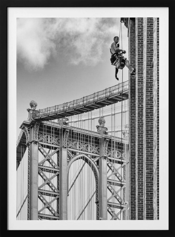 A black and white photograph capturing a window washer rappelling down the side of a tall brick building. In the background, the intricate steel structure of the Manhattan Bridge stands against a partly cloudy sky, creating a powerful urban scene. Decor