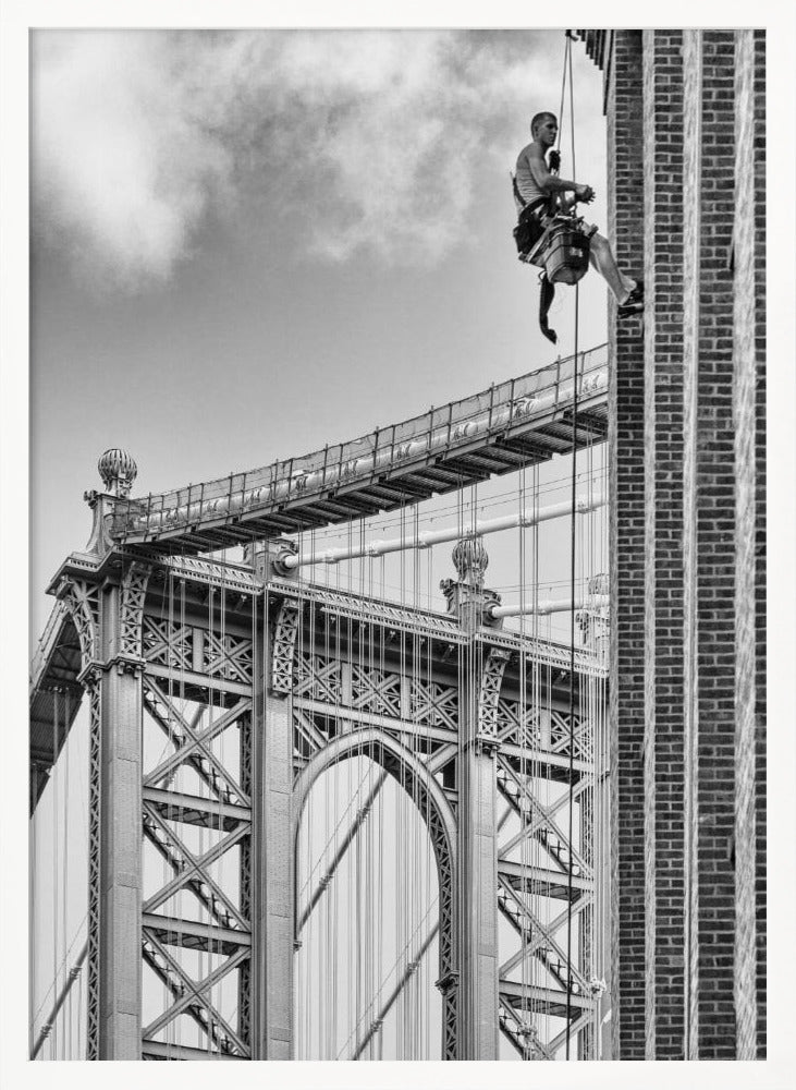 A black and white photograph capturing a window washer rappelling down the side of a tall brick building. In the background, the intricate steel structure of the Manhattan Bridge stands against a partly cloudy sky, creating a powerful urban scene. Decor