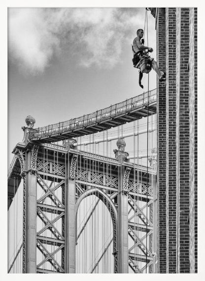 A black and white photograph capturing a window washer rappelling down the side of a tall brick building. In the background, the intricate steel structure of the Manhattan Bridge stands against a partly cloudy sky, creating a powerful urban scene. Decor