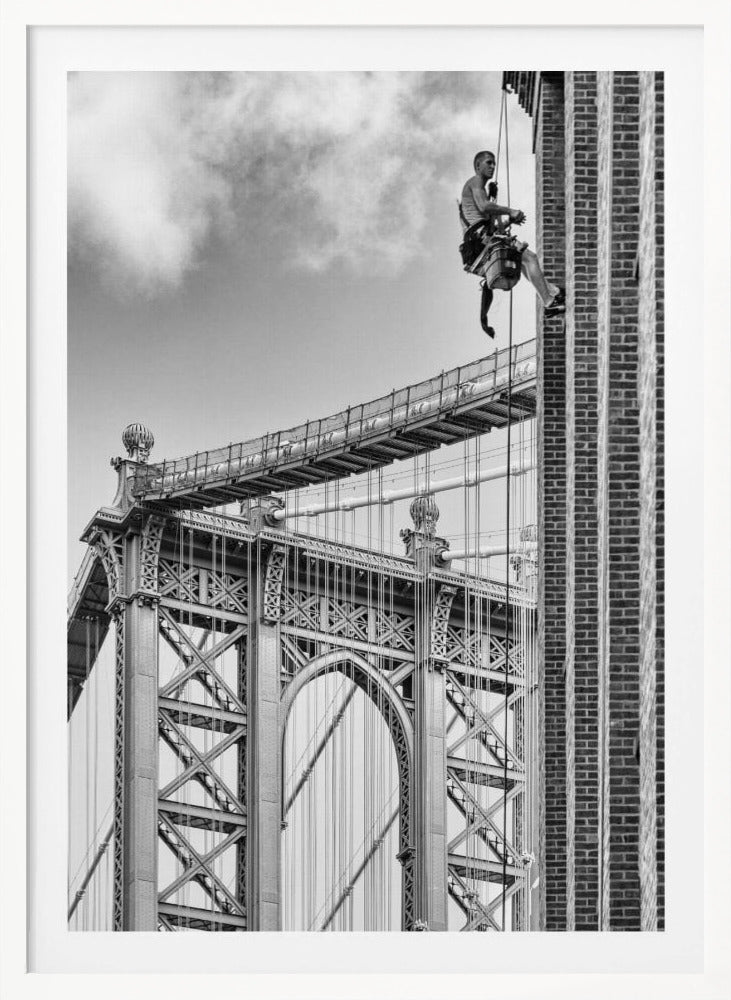 A black and white photograph capturing a window washer rappelling down the side of a tall brick building. In the background, the intricate steel structure of the Manhattan Bridge stands against a partly cloudy sky, creating a powerful urban scene. Decor