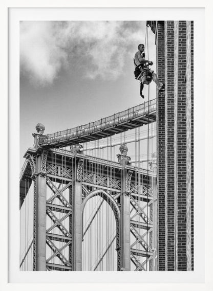 A black and white photograph capturing a window washer rappelling down the side of a tall brick building. In the background, the intricate steel structure of the Manhattan Bridge stands against a partly cloudy sky, creating a powerful urban scene. Decor