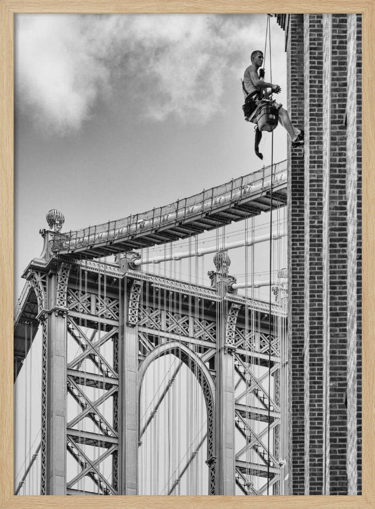 A black and white photograph capturing a window washer rappelling down the side of a tall brick building. In the background, the intricate steel structure of the Manhattan Bridge stands against a partly cloudy sky, creating a powerful urban scene. Decor