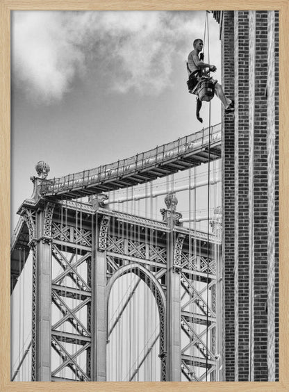 A black and white photograph capturing a window washer rappelling down the side of a tall brick building. In the background, the intricate steel structure of the Manhattan Bridge stands against a partly cloudy sky, creating a powerful urban scene. Decor