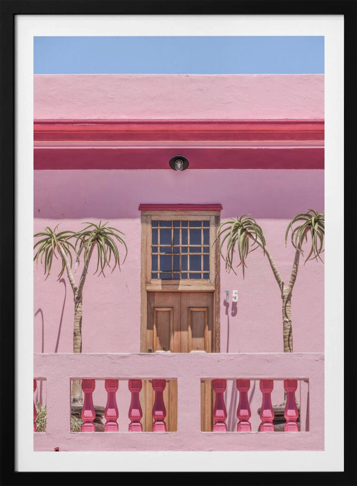 A brightly colored pink building stands under a clear blue sky, framed in black. A central wooden door with a paned window is flanked by two slender, palm-like trees. In the foreground, a pink balcony wall features fuchsia-colored balusters. Decor