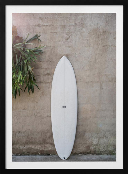 A minimalist photograph of a white surfboard leaning against a textured beige concrete wall. To the left, a large green staghorn fern adds a touch of nature. The entire scene is enclosed in a simple black frame. Artwork
