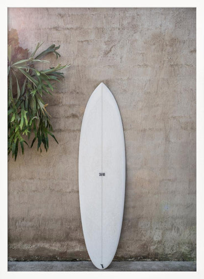 A minimalist photograph of a white surfboard leaning against a textured beige concrete wall. To the left, a large green staghorn fern adds a touch of nature. The entire scene is enclosed in a simple black frame. Artwork