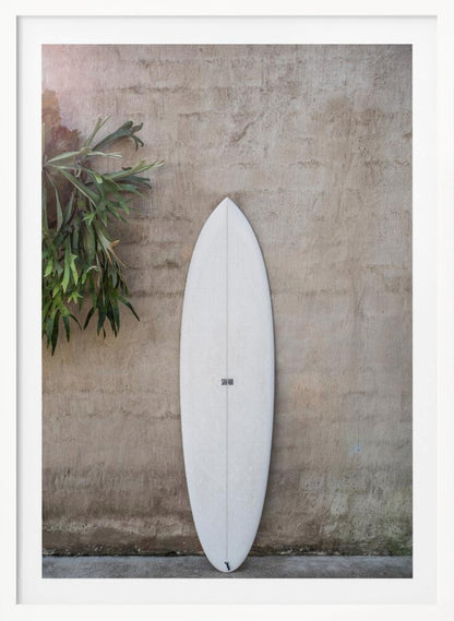 A minimalist photograph of a white surfboard leaning against a textured beige concrete wall. To the left, a large green staghorn fern adds a touch of nature. The entire scene is enclosed in a simple black frame. Artwork