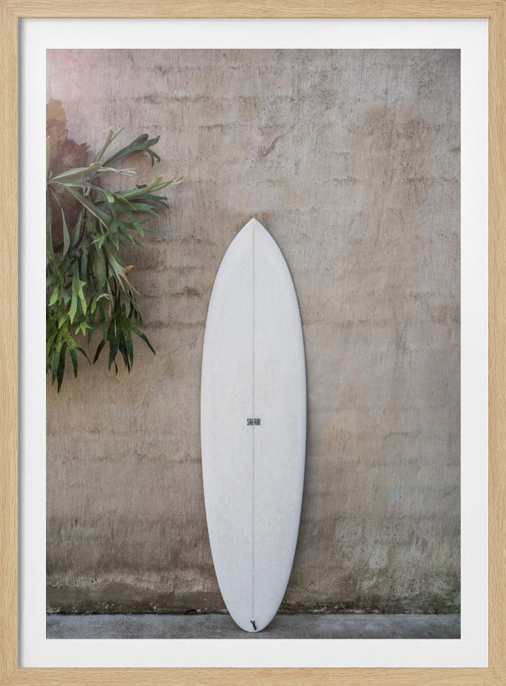 A minimalist photograph of a white surfboard leaning against a textured beige concrete wall. To the left, a large green staghorn fern adds a touch of nature. The entire scene is enclosed in a simple black frame. Artwork