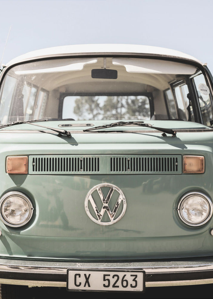 A close-up front view of a vintage sage green Volkswagen bus with a white roof. The iconic VW logo is on the front, with round headlights and a European license plate, set against a pale blue sky. Print