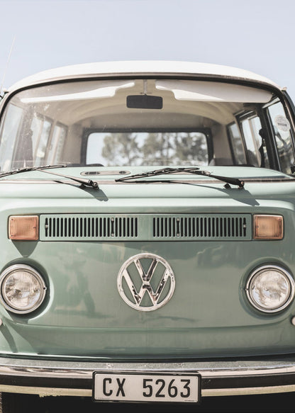 A close-up front view of a vintage sage green Volkswagen bus with a white roof. The iconic VW logo is on the front, with round headlights and a European license plate, set against a pale blue sky. Print