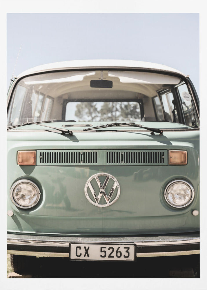 A close-up front view of a vintage sage green Volkswagen bus with a white roof. The iconic VW logo is on the front, with round headlights and a European license plate, set against a pale blue sky. Print