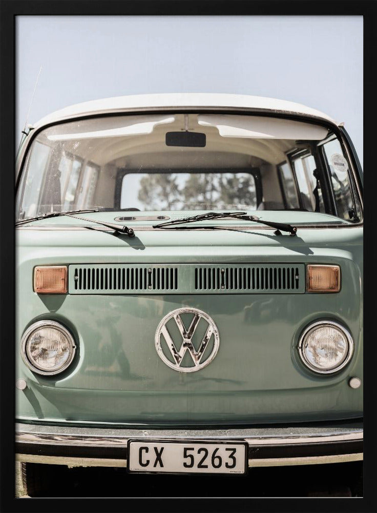 A close-up front view of a vintage sage green Volkswagen bus with a white roof. The iconic VW logo is on the front, with round headlights and a European license plate, set against a pale blue sky. Print