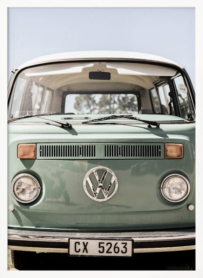 A close-up front view of a vintage sage green Volkswagen bus with a white roof. The iconic VW logo is on the front, with round headlights and a European license plate, set against a pale blue sky. Print