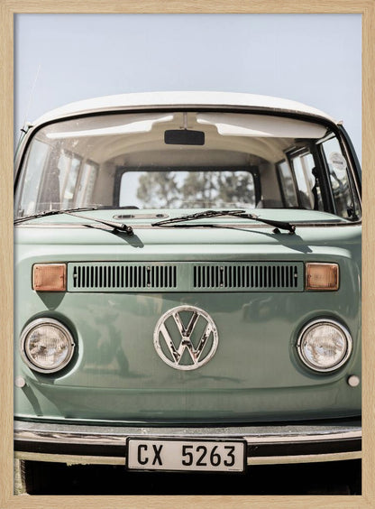 A close-up front view of a vintage sage green Volkswagen bus with a white roof. The iconic VW logo is on the front, with round headlights and a European license plate, set against a pale blue sky. Print