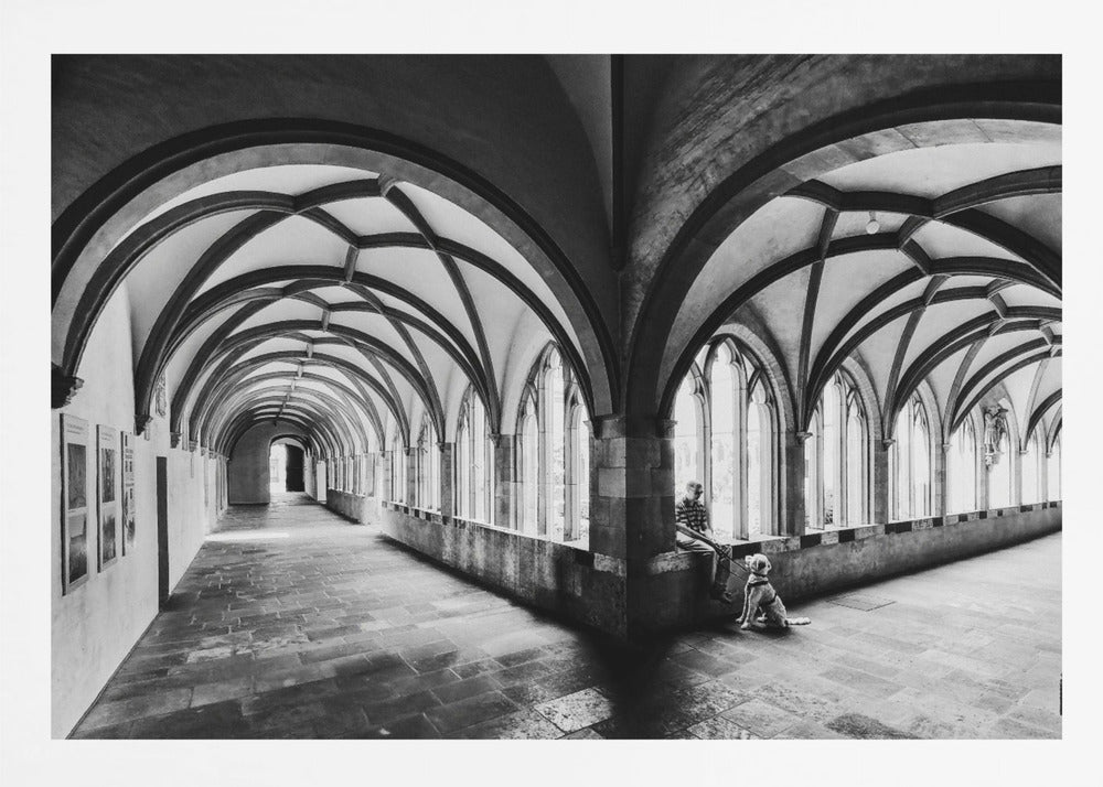 A black and white photograph of a person and their dog sitting in the corner of a Gothic cloister. The stone hallway features a dramatic vaulted ceiling and a long row of arched windows, creating a powerful sense of perspective and light. Decor