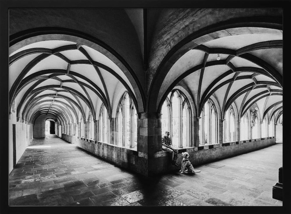A black and white photograph of a person and their dog sitting in the corner of a Gothic cloister. The stone hallway features a dramatic vaulted ceiling and a long row of arched windows, creating a powerful sense of perspective and light. Decor