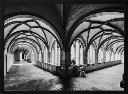 A black and white photograph of a person and their dog sitting in the corner of a Gothic cloister. The stone hallway features a dramatic vaulted ceiling and a long row of arched windows, creating a powerful sense of perspective and light. Decor