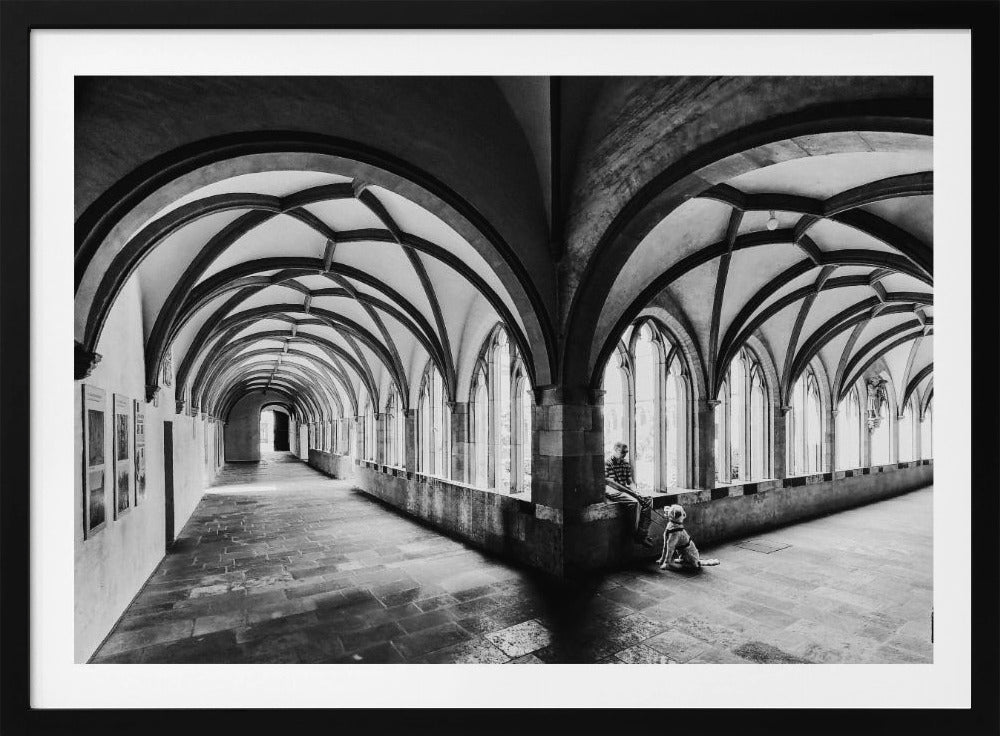 A black and white photograph of a person and their dog sitting in the corner of a Gothic cloister. The stone hallway features a dramatic vaulted ceiling and a long row of arched windows, creating a powerful sense of perspective and light. Decor
