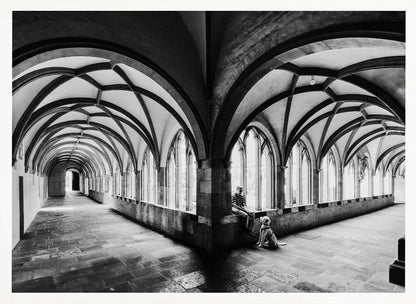 A black and white photograph of a person and their dog sitting in the corner of a Gothic cloister. The stone hallway features a dramatic vaulted ceiling and a long row of arched windows, creating a powerful sense of perspective and light. Decor