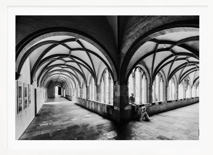 A black and white photograph of a person and their dog sitting in the corner of a Gothic cloister. The stone hallway features a dramatic vaulted ceiling and a long row of arched windows, creating a powerful sense of perspective and light. Decor