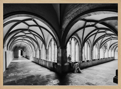 A black and white photograph of a person and their dog sitting in the corner of a Gothic cloister. The stone hallway features a dramatic vaulted ceiling and a long row of arched windows, creating a powerful sense of perspective and light. Decor