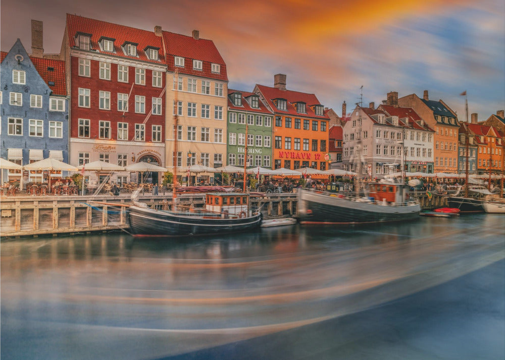 A long-exposure photograph of the colorful Nyhavn harbor in Copenhagen at sunset. Historic, brightly colored buildings line the canal, where two traditional wooden boats are moored. The sky is a dramatic blend of orange and blue, and the water is blurred, showing streaks of reflected light. Decor