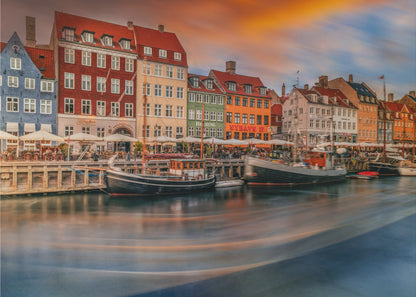 A long-exposure photograph of the colorful Nyhavn harbor in Copenhagen at sunset. Historic, brightly colored buildings line the canal, where two traditional wooden boats are moored. The sky is a dramatic blend of orange and blue, and the water is blurred, showing streaks of reflected light. Decor