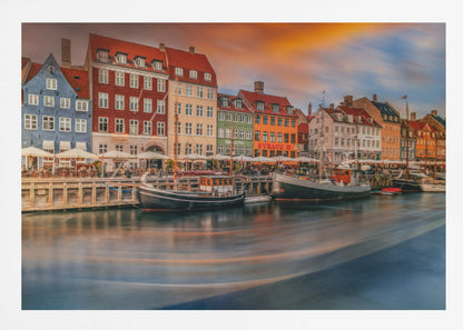 A long-exposure photograph of the colorful Nyhavn harbor in Copenhagen at sunset. Historic, brightly colored buildings line the canal, where two traditional wooden boats are moored. The sky is a dramatic blend of orange and blue, and the water is blurred, showing streaks of reflected light. Decor