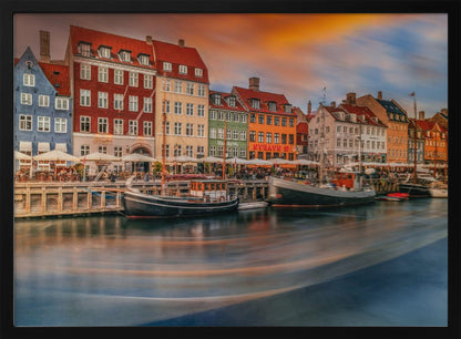 A long-exposure photograph of the colorful Nyhavn harbor in Copenhagen at sunset. Historic, brightly colored buildings line the canal, where two traditional wooden boats are moored. The sky is a dramatic blend of orange and blue, and the water is blurred, showing streaks of reflected light. Decor